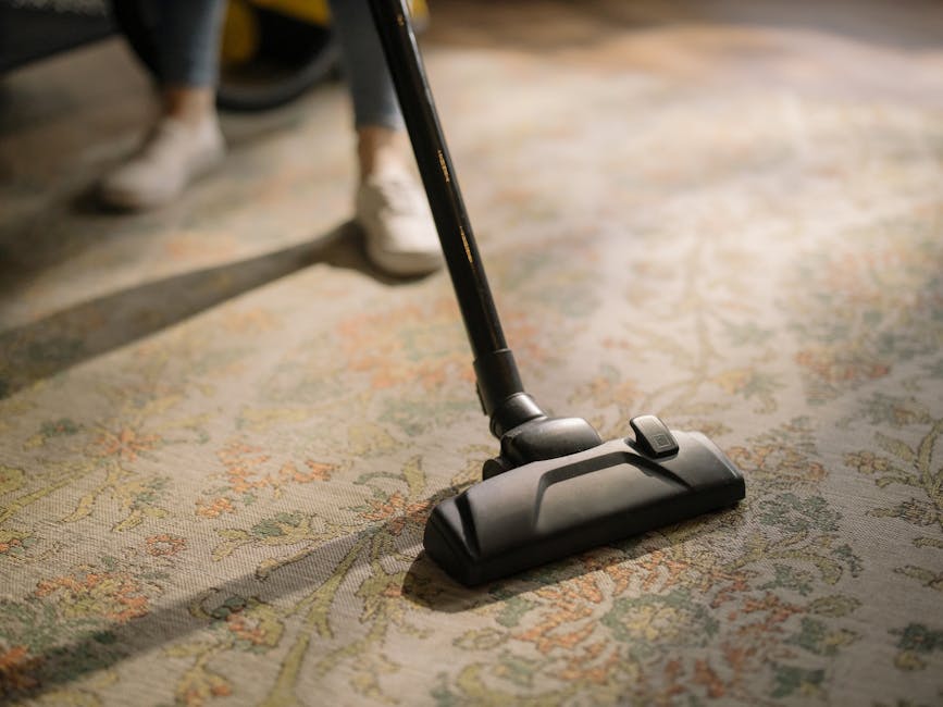 A person in grey overalls is engaged in surface cleaning using a yellow professional vacuum cleaner in a room with dark wall panels and a white beam above. The individual is bent over, focused on the 