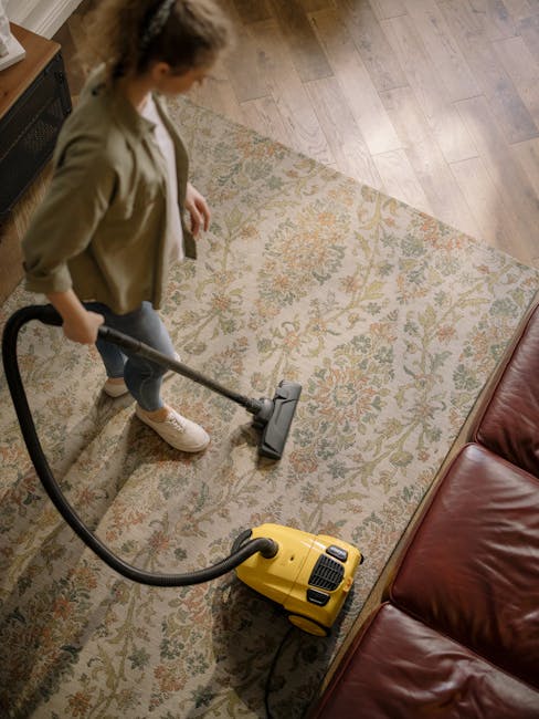 A close-up of a vacuum cleaner attachment being used to clean a patterned carpet in a residential living room. The carpet features intricate floral designs in muted colors and appears clean and well-m
