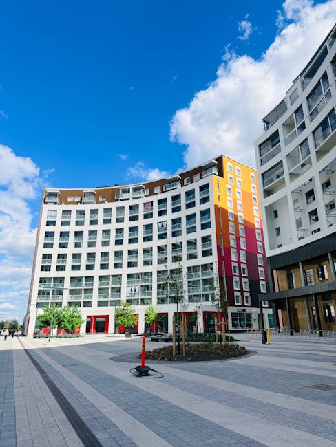 A modern multi-story residential building with a curved facade, featuring large glass windows and white exterior walls accented by orange, pink, and yellow panels. The building is situated in a spacio