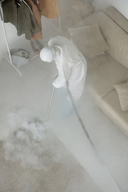 A professional cleaner dressed in white protective coveralls, wearing an face mask and orange gloves, is operating a vacuum or cleaning device on a smooth, grey tiled floor in a modern residential int