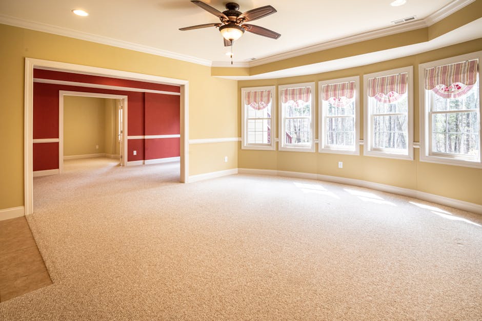 A spacious, well-lit living room with cream carpet flooring, featuring a large bay window with five sections adorned with pink and white striped valances. The walls are painted in a soft yellow, accen