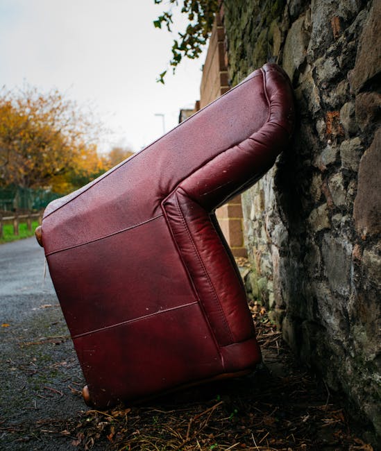 A red leather upholstered armchair with visible seams and padding, positioned outdoors on a damp pavement next to a rough stone wall. The chair is tilted and appears to be discarded or abandoned, with
