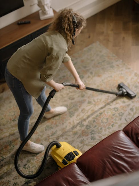 A professional cleaning technician from Ilford Carpet Cleaners is performing deep cleaning in a residential living room, wearing full protective gear including a white suit, gloves, and a face mask. T